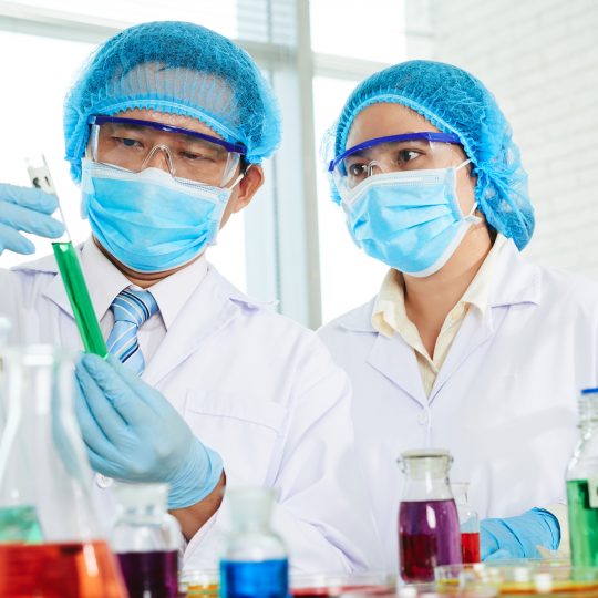 Concentrated Asian scientists wearing white coats and medical masks examining liquid in test tube while gathered together at modern laboratory